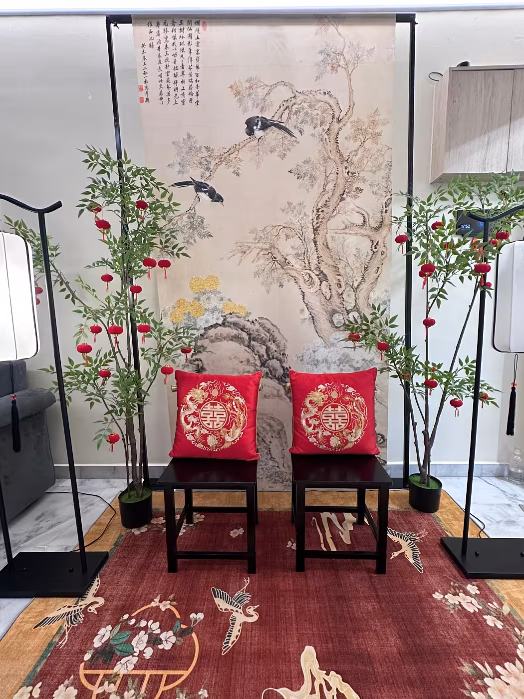 A traditional Chinese wedding tea ceremony vignette centred on two dark wooden chairs dressed with embroidered red dragon and phoenix double-happiness cushion covers, set against an ancient cypress ink painting hanging cloth framed by a black metal arch. Flanking artificial green nandina trees strung with miniature red velvet lanterns and a vibrant red crane-motif floor carpet complete the auspicious, richly layered scene.