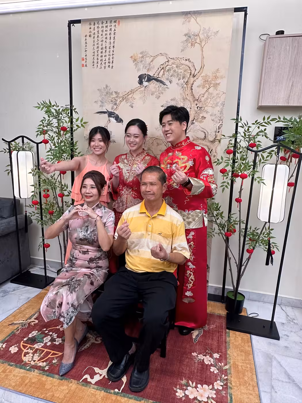 A traditional Chinese tea ceremony setup anchored by an ancient green cypress double happiness ink painting backdrop on a black metal arch stand, flanked by artificial potted nandina trees adorned with small red velvet hanging lanterns and retro arch vertical LED floor lamps, all grounded on a vibrant red floral carpet.