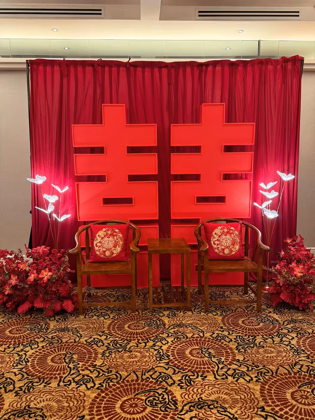 A bold red Chinese wedding vignette centres on a large 囍 Double Happiness backdrop draped with rich pleated crimson curtain fabric, flanked by glowing 5-head butterfly LED walkway lights and lush red artificial floor flowers. A rustic walnut wooden three-piece chair-and-table set, dressed with embroidered dragon-and-phoenix cushion covers, anchors the elegant traditional setup.
