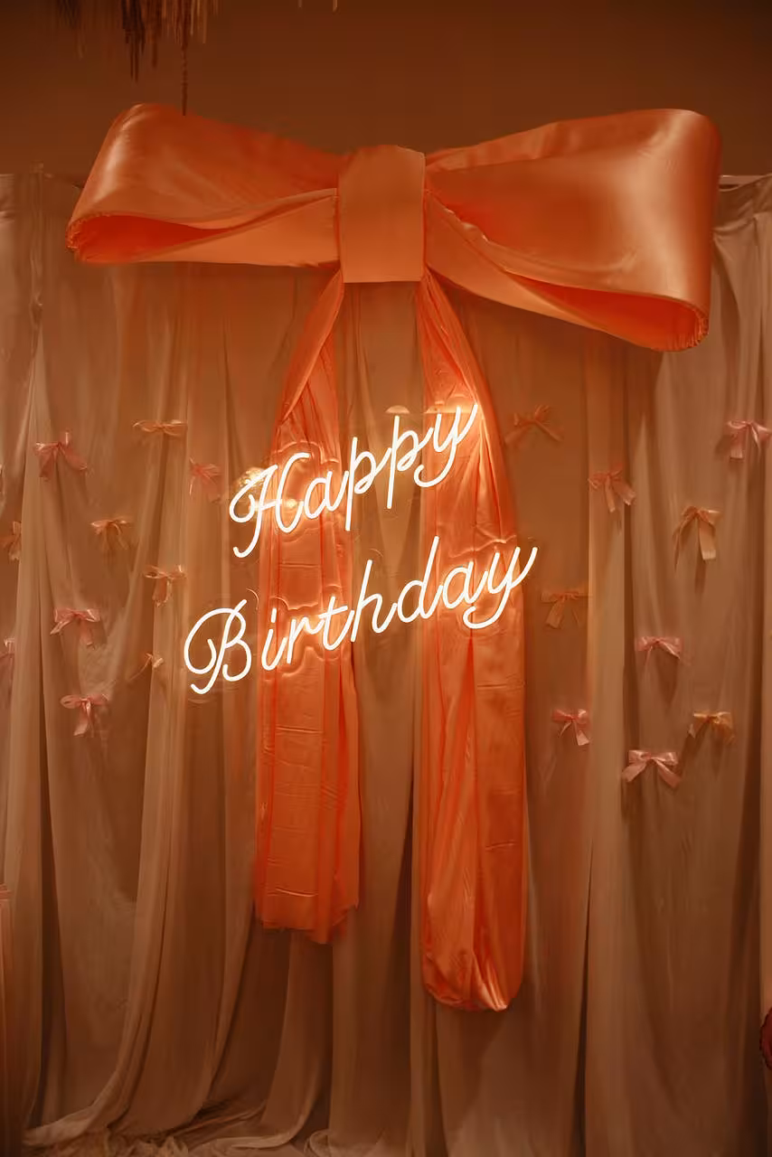 A glowing white Happy Birthday LED Neon Light Signage in flowing cursive script takes centre stage against the draped Wedding Fabric Joyous White Celebration Curtain, adorned with scattered small pink ribbon bows. A voluminous peach satin bow crowns the backdrop, creating a warm, romantic birthday atmosphere.