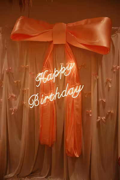 Happy Birthday LED neon sign glowing in cursive against a white fabric curtain backdrop with a large peach satin bow