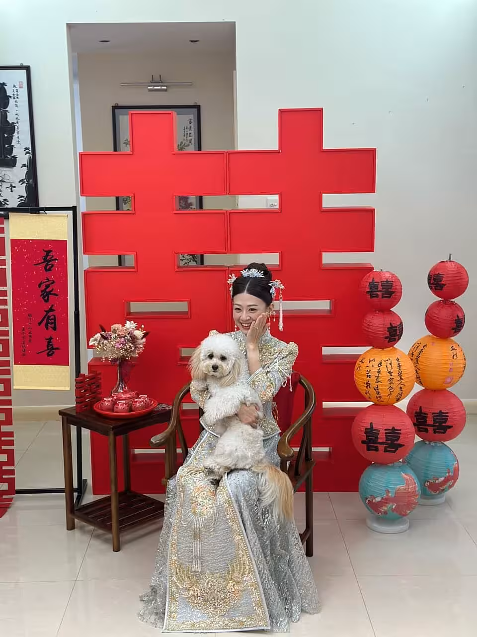 A striking 2-metre red 囍 Double Happiness backdrop anchors the scene, paired with a tower of stacked 喜-printed lanterns in vibrant red, orange, and teal. A rustic walnut wooden chair sits in the foreground beside a side table dressed with a pink dried floral arrangement and a red ceramic tea set, completed by a calligraphy scroll poster to the left.
