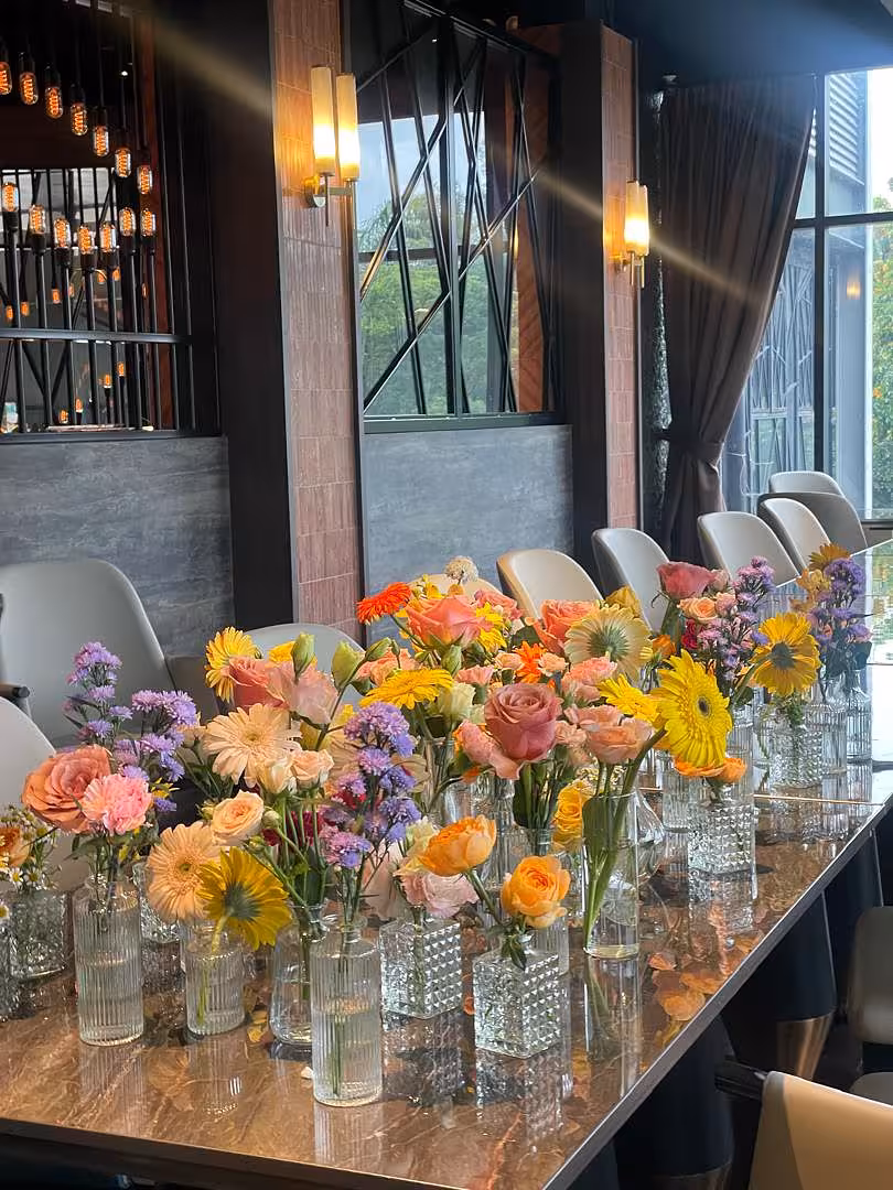 A lush tablescape of Mini Clear Glass Flower Vases in Types A, B, C, and D lines a long marble table, each holding vibrant clusters of yellow gerberas, peach and pink roses, purple statice, and orange blooms. The mix of ribbed, smooth, and textured glass silhouettes adds dimension, while the warm ambient lighting of the venue deepens the cheerful, garden-fresh mood.