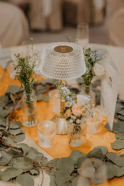 Crystal shade portable dining table lamp on a gold tray surrounded by clear glass bud vases with pastel florals, LED tealight candle cups, and trailing eucalyptus greenery