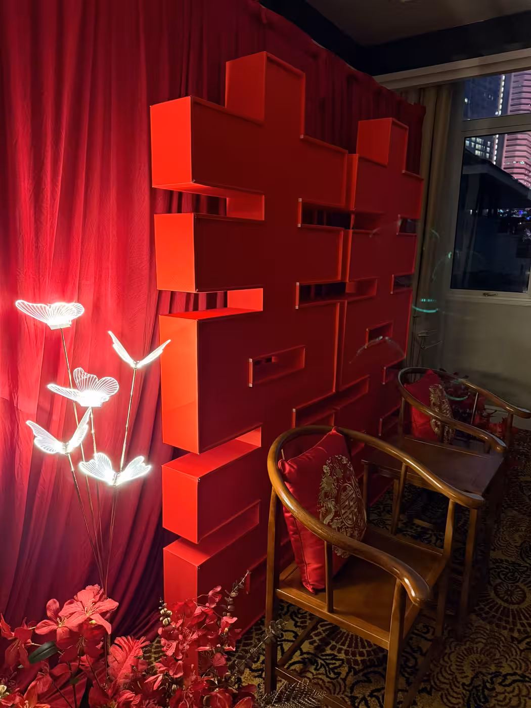 A rich all-red Chinese wedding vignette anchored by a dramatic pleated fabric curtain and a towering 3D 囍 Double Happiness backdrop. Glowing white butterfly LED lights and lush red artificial blooms frame the left side, while rustic walnut round-back chairs dressed with embroidered dragon and phoenix cushions complete the traditional setting.