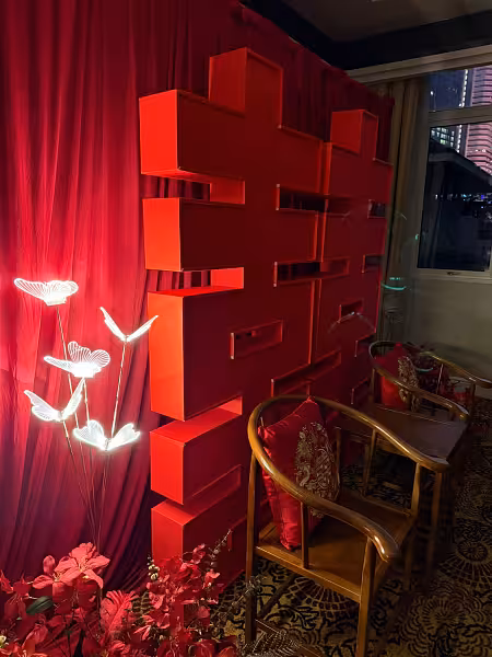 Red pleated curtain backdrop with 3D 囍 Double Happiness structure, glowing butterfly LED walkway lights, red artificial floor flowers, and rustic walnut chairs with dragon and phoenix embroidered cushions
