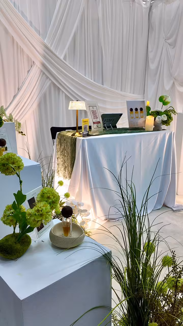 Flowing ivory drape fabric sweeps dramatically across the backdrop, setting a soft, elegant tone behind a white-clothed table styled with a crumpled sage-green runner, a warm-glowing crystal portable lamp, and lush green artificial floral arrangements in a textured pedestal bowl vase.