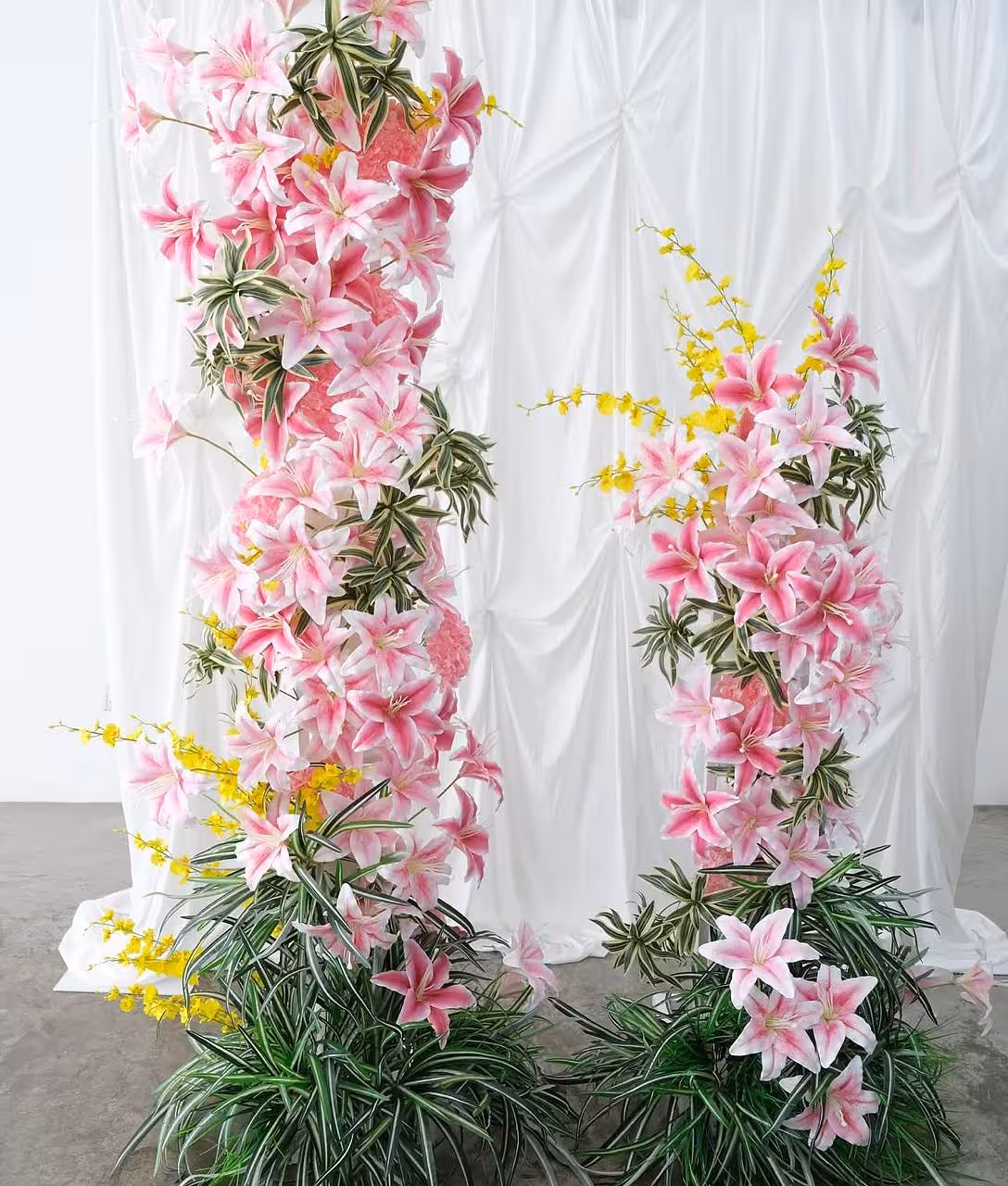 Two lush artificial flower arch columns densely layered with pink stargazer lilies, delicate yellow blossom sprays, and variegated green foliage cascade floor-to-ceiling against a softly ruched white fabric backdrop, creating a romantic and vibrant celebration setting.