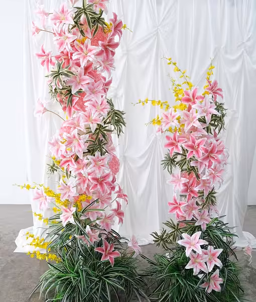 Two tall pink artificial lily flower arch columns with yellow sprays and lush green foliage against a white draped fabric backdrop