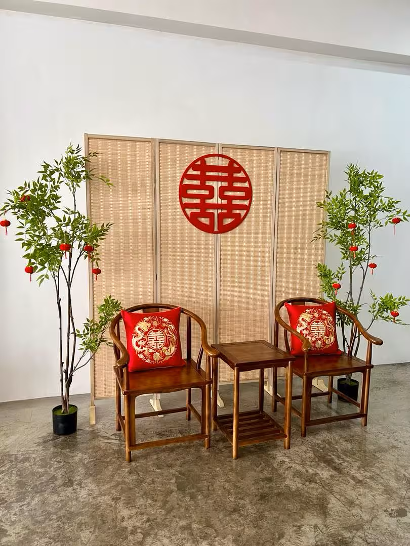A four-panel bamboo folding partition forms a warm natural backdrop, anchored by a bold red double-happiness wall ornament at its centre. Flanking artificial nandina trees adorned with small hanging red velvet lanterns frame a rustic walnut wooden chair-and-table set, whose seats are dressed with richly embroidered red dragon-and-phoenix cushion covers.