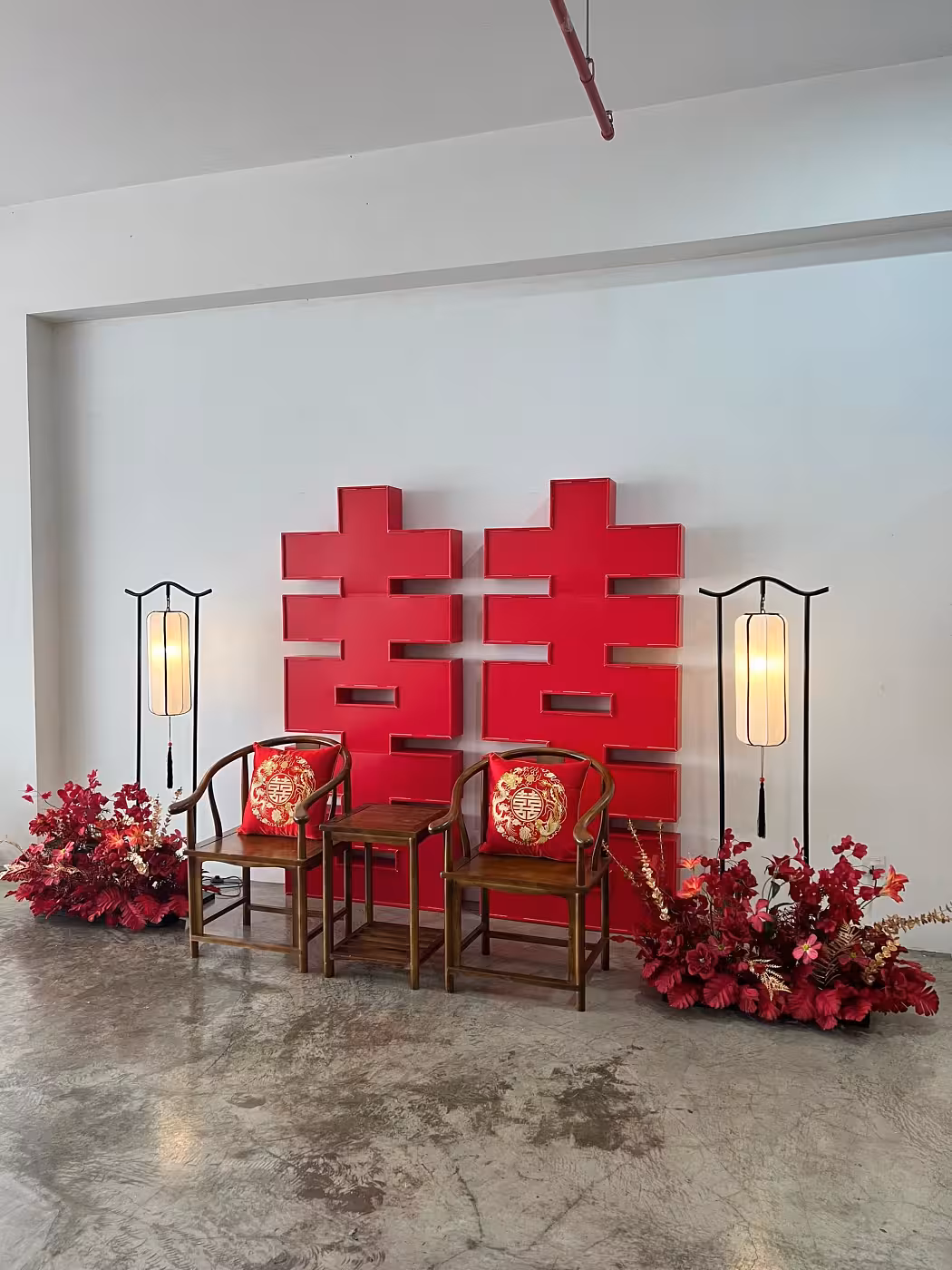A striking 2-metre bold red 囍 Double Happiness backdrop anchors this traditional Chinese-inspired setup, framed by a pair of glowing Retro Arch Vertical Lantern LED Floor Lamps in black iron. Two rustic walnut horseshoe chairs dressed with gold-embroidered dragon and phoenix cushions sit at the centre, while lush deep red artificial floor flower arrangements ground each side of the scene.