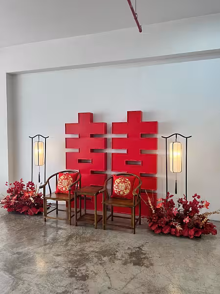 Bold red 囍 Double Happiness backdrop flanked by arch lantern floor lamps and rustic walnut chairs with embroidered red cushions and artificial floral arrangements