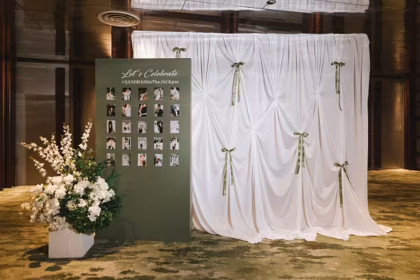 White celebration curtain backdrop with sage green ribbon bows beside a sage green photo display board and white floral arrangement