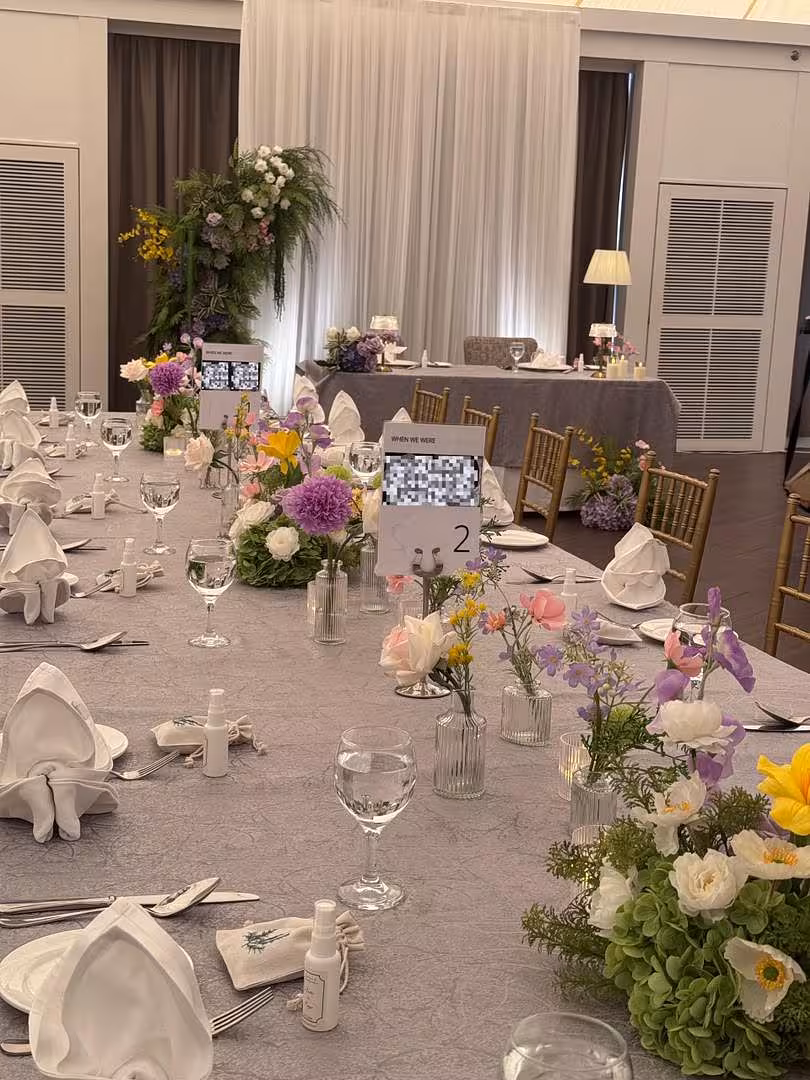 A grey linen-draped banquet table is styled with an abundant mix of wildflowers — including purple dahlias, pink roses, yellow blooms, and lavender stems — arranged in assorted Mini Clear Glass Flower Vases (Types A, B, C, and D) clustered along the centre. A dramatic tall floral installation of greenery, purple, and white flowers anchors the backdrop behind sheer white draping, tying the garden-inspired palette together.