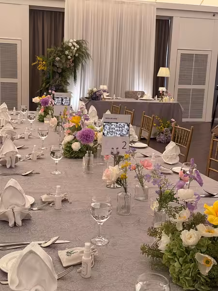 Round banquet table with mixed wildflowers in mini clear glass vases of varying shapes, grey linen, folded napkins, and a tall floral backdrop arrangement