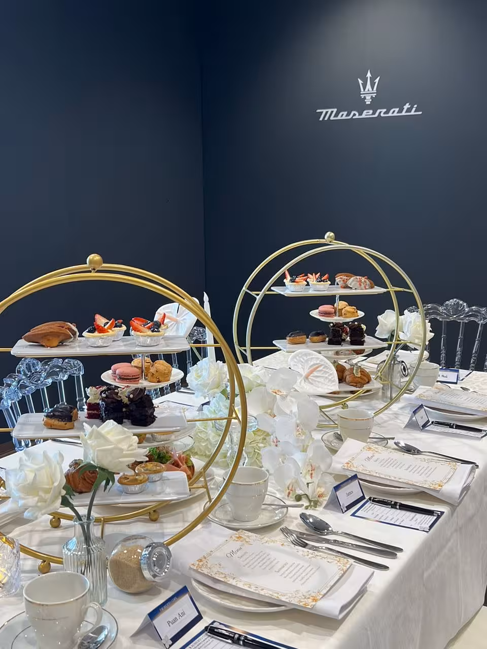 Two gold round 3-layer dessert display racks anchor this elegant navy-and-white tablescape, surrounded by lush white artificial floral arrangements and glass pedestal taper candle holders with slim LED candles. Crisp white table linens, printed menu cards, and name place cards complete the refined, luxurious setting.