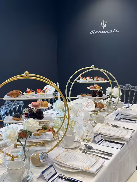 Two gold round 3-layer dessert display racks styled with white artificial flowers and taper candles on a white-draped table against a navy wall