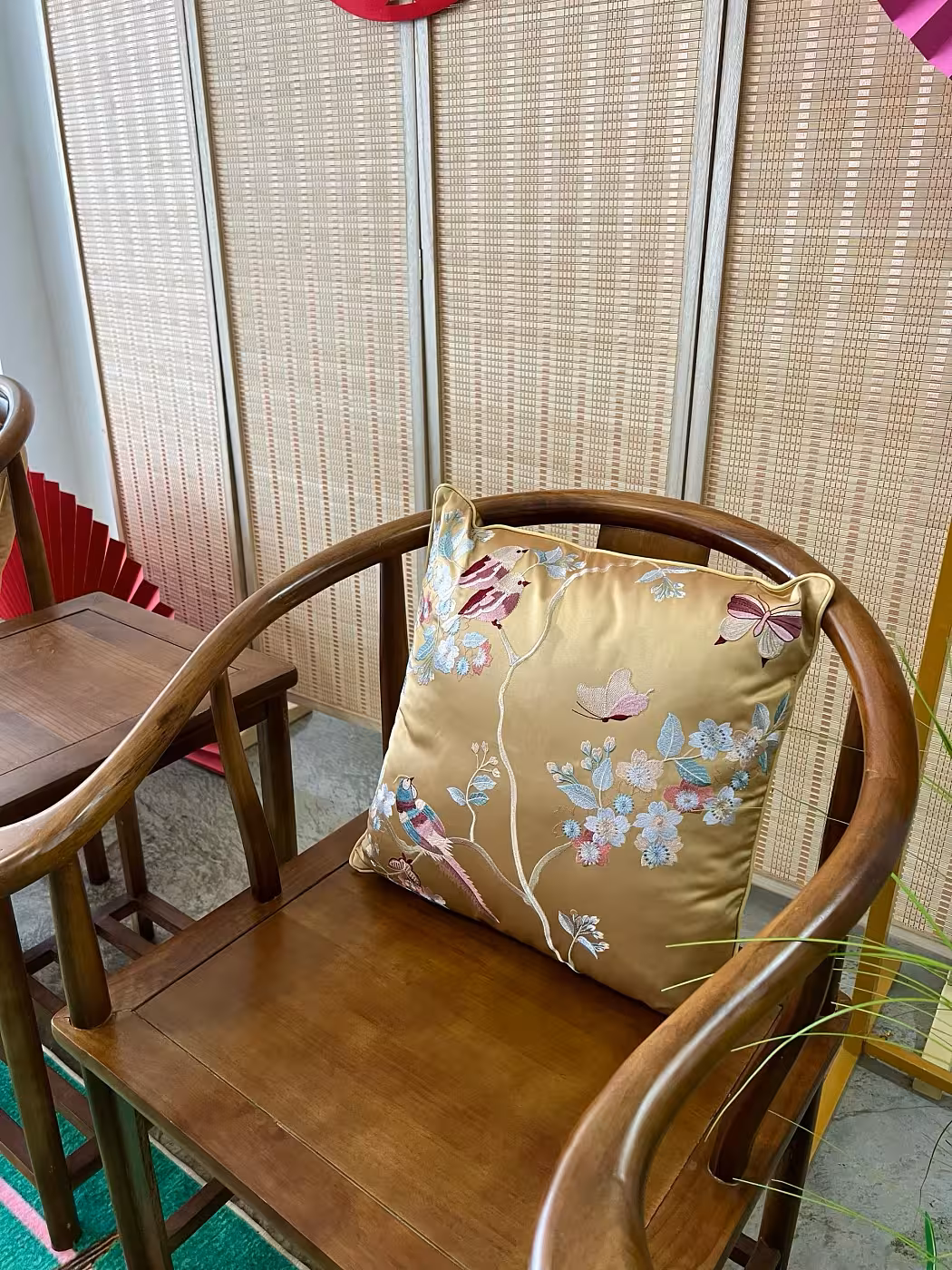A luxurious Oriental Embroidered Floral Bird Butterfly Designed Decorative Satin Cushion Cover in gold takes centre stage, its intricate embroidery featuring delicate blooms, perched birds, and butterflies in soft pink, blue, and white. The setting is completed by a rich dark wood armchair, a woven bamboo panel backdrop, and glimpses of red paper décor accents.