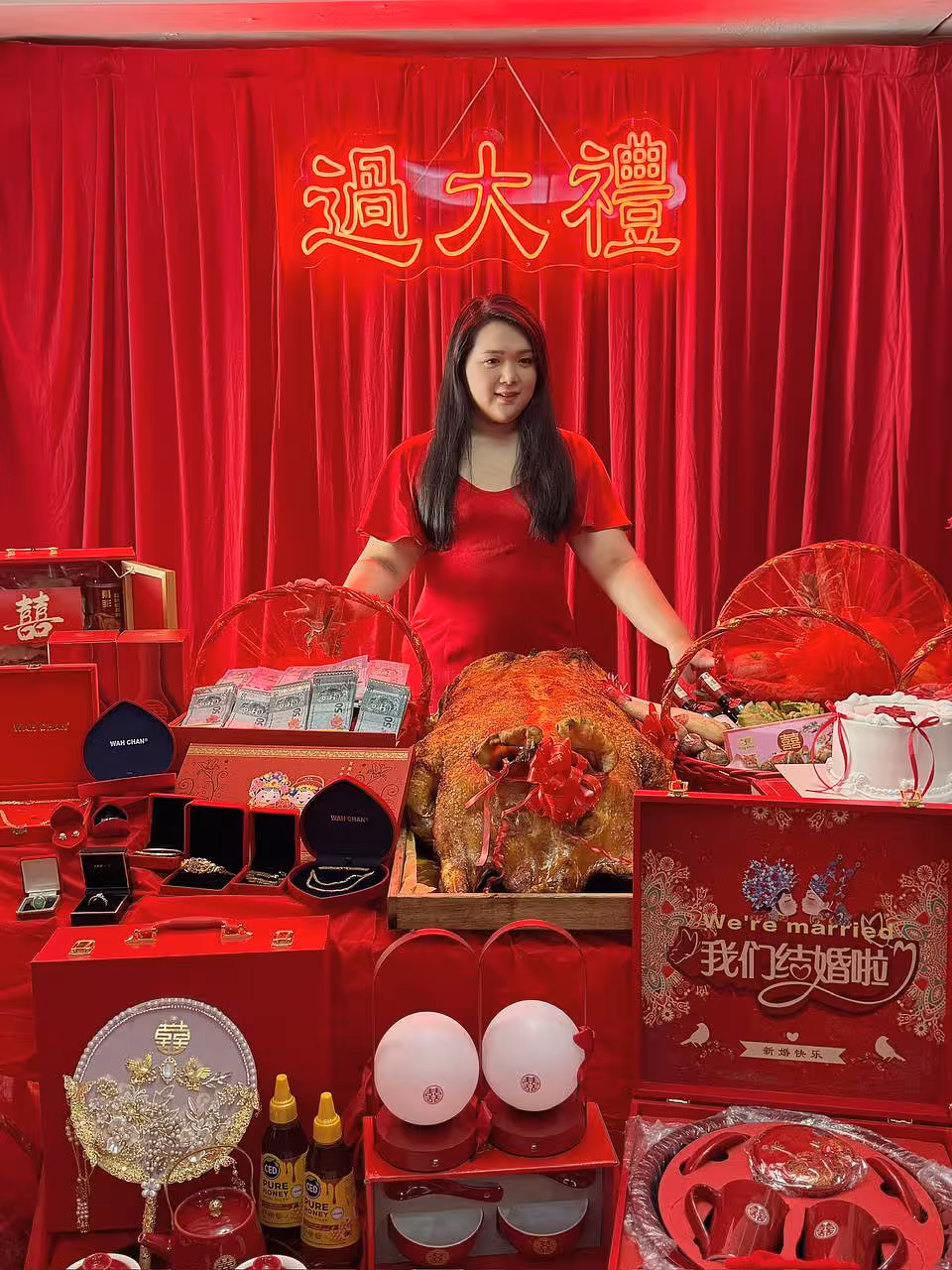 A bold red velvet curtain backdrop frames the glowing 過大禮 GuoDaLi neon light in warm gold, casting a festive glow over the styled display below. The table is richly arranged with traditional betrothal gifts including jewellery boxes, red gift baskets, a roasted pig, decorative fans, and auspicious double-happiness motif accessories, all unified in a vivid red-and-gold colour palette.