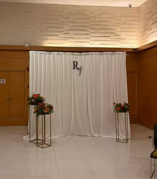 White drape fabric backdrop with couple's monogram initial sign flanked by two gold iron stands topped with orange floral arrangements