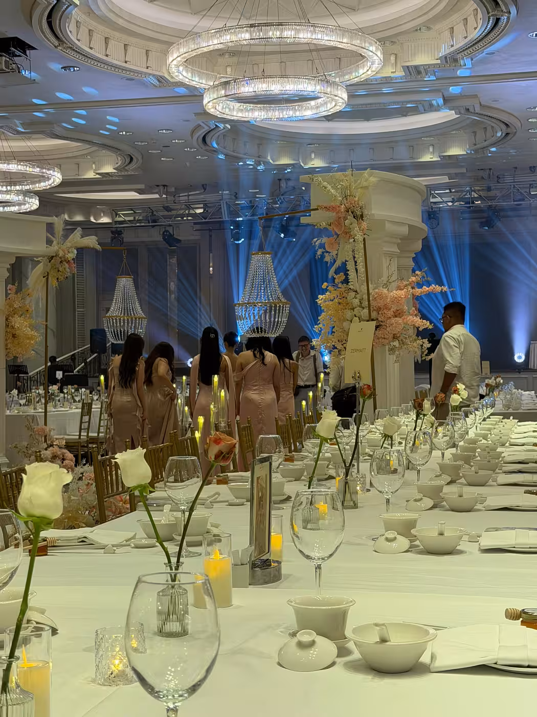 Long banquet tables dressed in crisp white linens are dotted with Mini Clear Glass Flower Vases (Type C) holding single-stem roses and LED Flickering Flameless Acrylic Candles casting a warm amber glow. Towering blush and white floral arrangements, crystal chandeliers overhead, and sweeping blue uplighting complete the luxurious ballroom atmosphere.