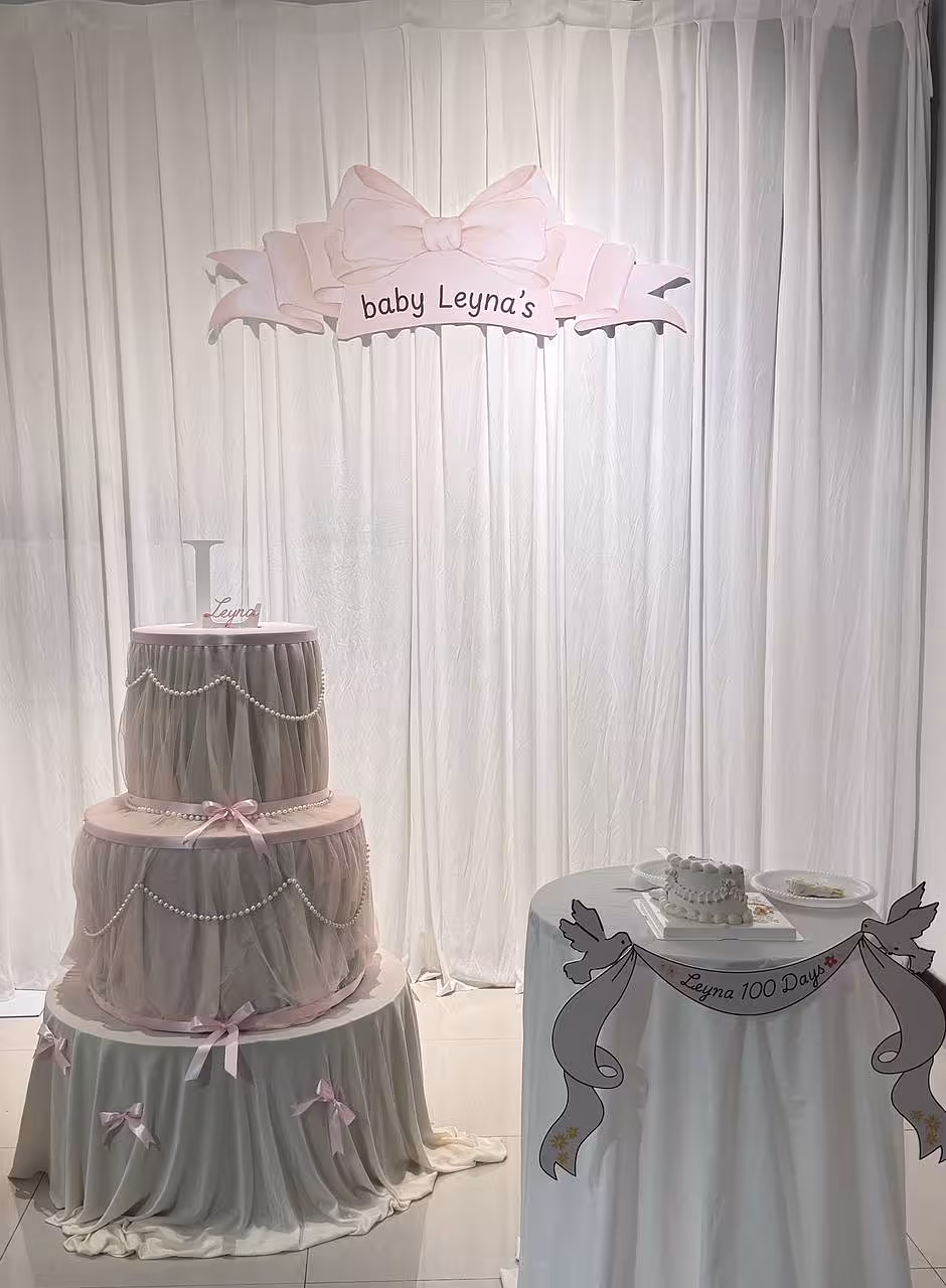 A dreamy soft pink and grey setup featuring a 3-Tier Cake Telescopic Stand layered with flowing grey tulle, pearl garlands, and pink satin bows, set against the elegant pleated folds of the Wedding Fabric Joyous White Celebration Curtain. A large pink bow-and-banner prop overhead adds a whimsical finishing touch to the styled corner.