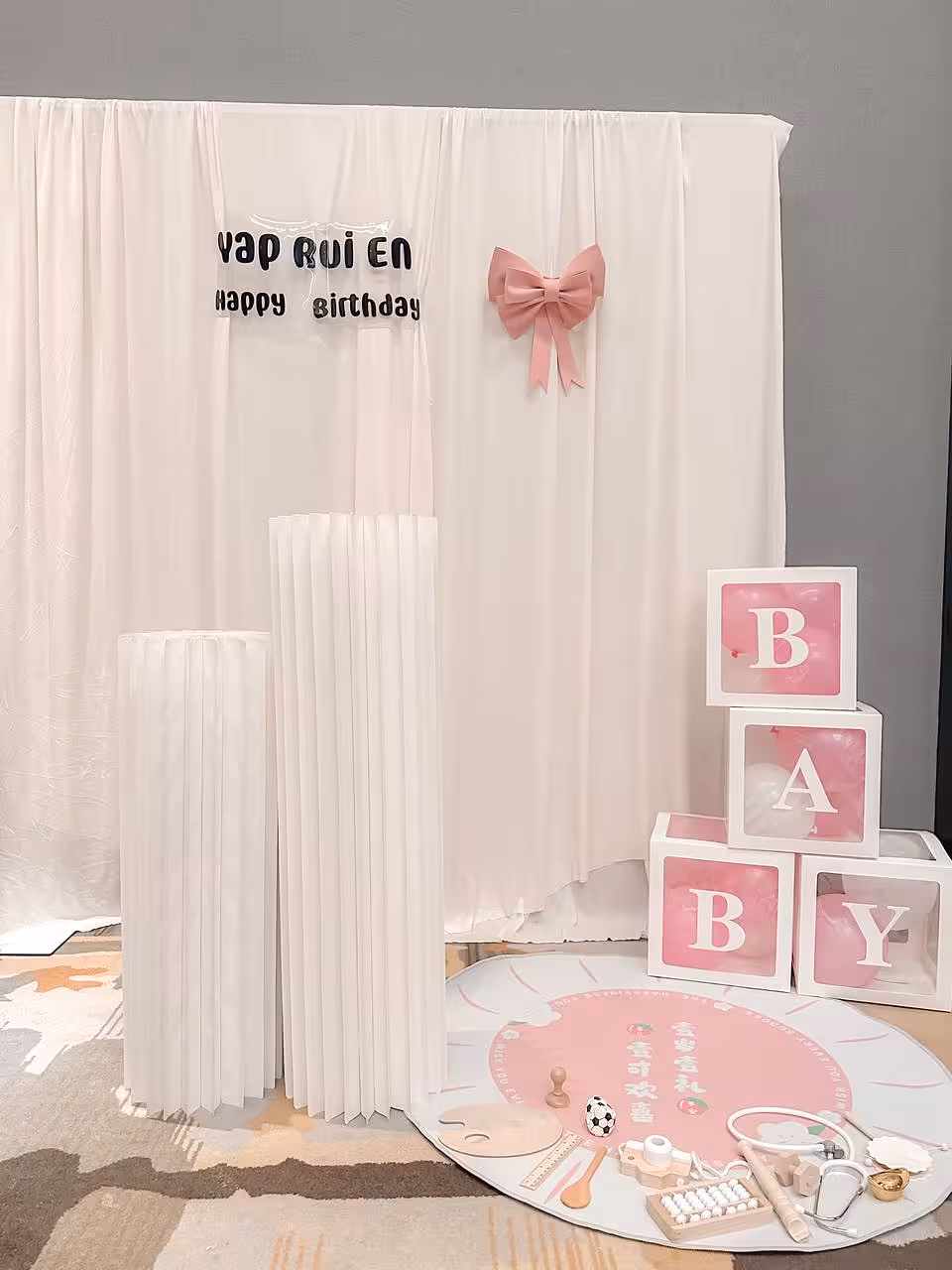 A soft champagne stretchable drape backdrop forms a full wall of gathered fabric, accented with a pink satin bow and a personalised birthday sign. Two white origami cylindrical pleated dessert stands anchor the foreground, complemented by stacked transparent BABY letter boxes filled with pink balloons for a sweet, pastel-toned celebration setup.