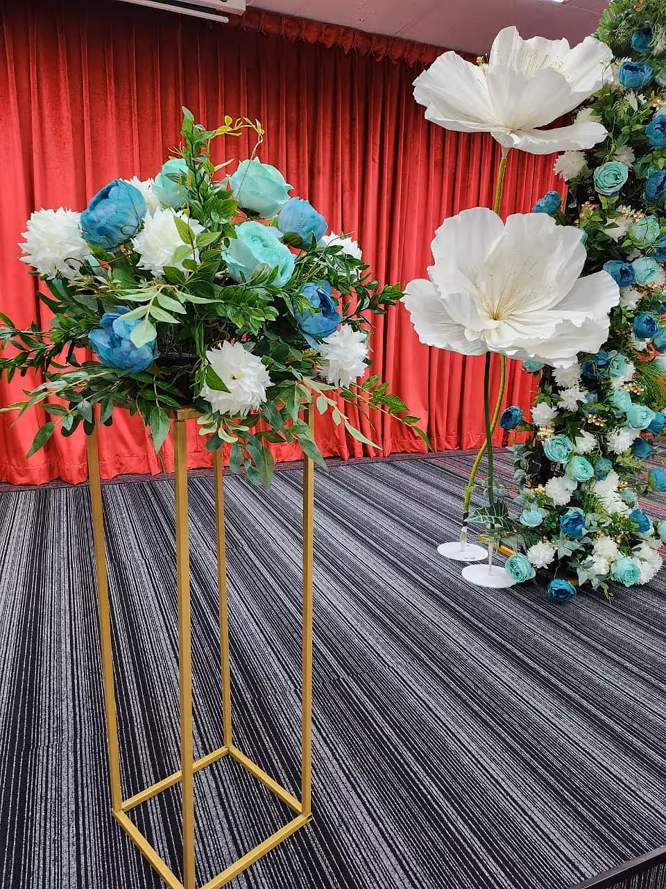 A tall Gold Iron Decorative Event Stand in rich gold metal supports a lush arrangement of teal, blue, and white blooms with trailing greenery, while a matching 80 cm stand nearby anchors oversized white paper flowers and a cascading floral arch — all set against deep red velvet draping.
