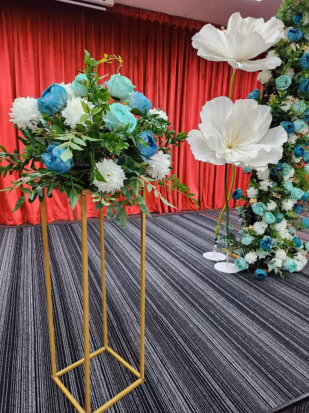Gold iron event stands topped with teal and white floral arrangement, beside a floral arch with oversized white blooms against a red curtain backdrop