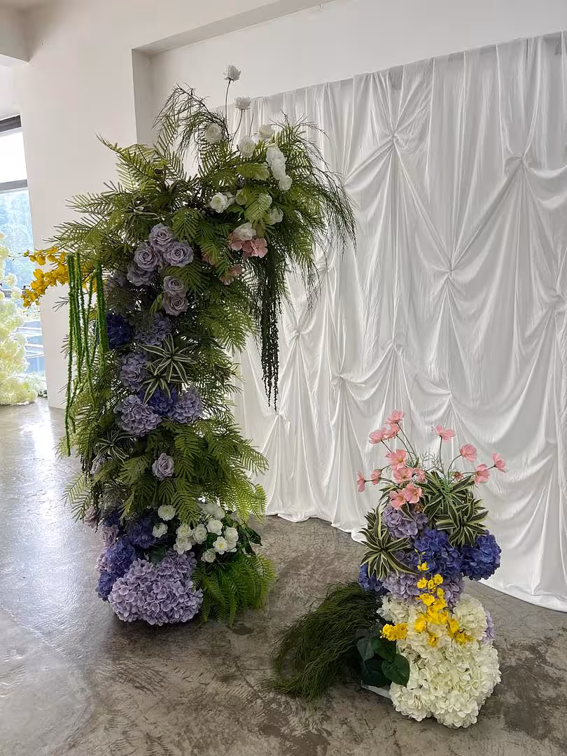 The Sweet Forest Artificial Flower Arch panels are densely layered with purple hydrangeas, lavender roses, pink cosmos, yellow blooms, and cascading fern-like greenery in a rich botanical style. Behind the arch, a softly ruched white celebration curtain forms a clean, elegant backdrop that beautifully offsets the vivid floral colour palette.
