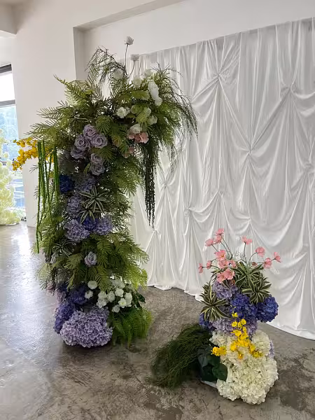 Sweet Forest artificial flower arch panels with purple hydrangeas, lavender roses, and lush greenery against a white pleated fabric backdrop
