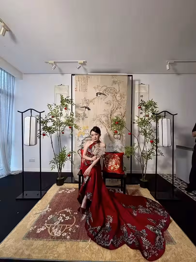 A traditional Chinese tea ceremony setting anchored by an Ancient Green Cypress Double Happiness ink painting hanging cloth, flanked by artificial Nandina potted plants and sleek black arch lantern floor lamps. A dragon and phoenix embroidered red cushion rests on a rustic wooden black copper chair, all grounded by a vibrant red floral carpet.