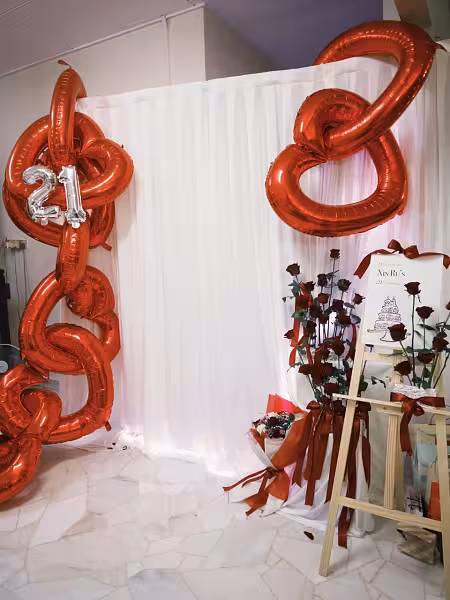 White fabric curtain backdrop with tall red foil balloon columns, a "21" silver balloon, red rose arrangements, and a wooden easel welcome sign with satin ribbon
