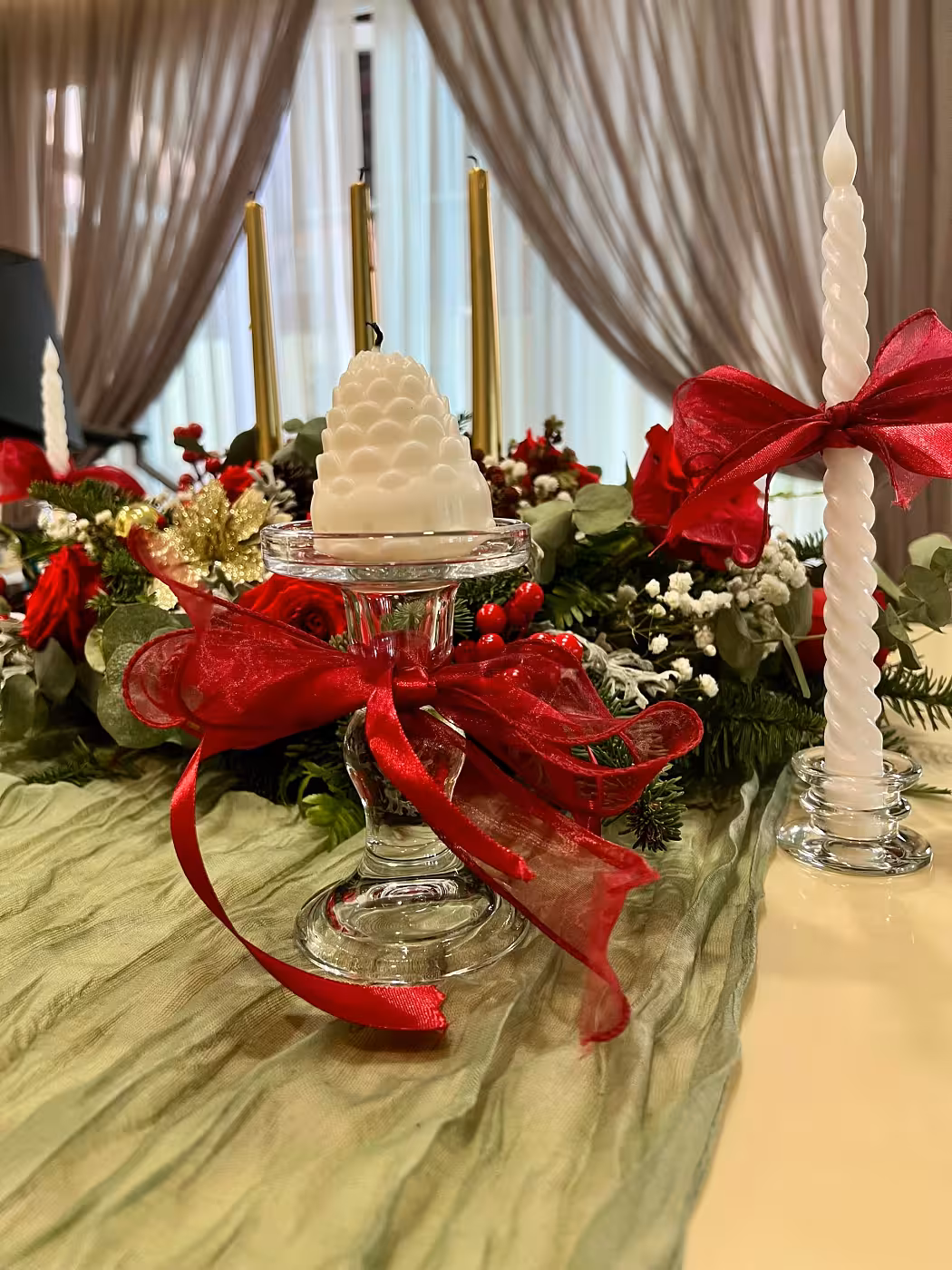 A crumpled sage table runner sets a soft, textural base for glass European pedestal candle holders dressed with bold red ribbon bows, a white LED spiral twisted taper candle, and a lush garland of red roses, pine sprigs, holly berries, and white blooms running along the centre.