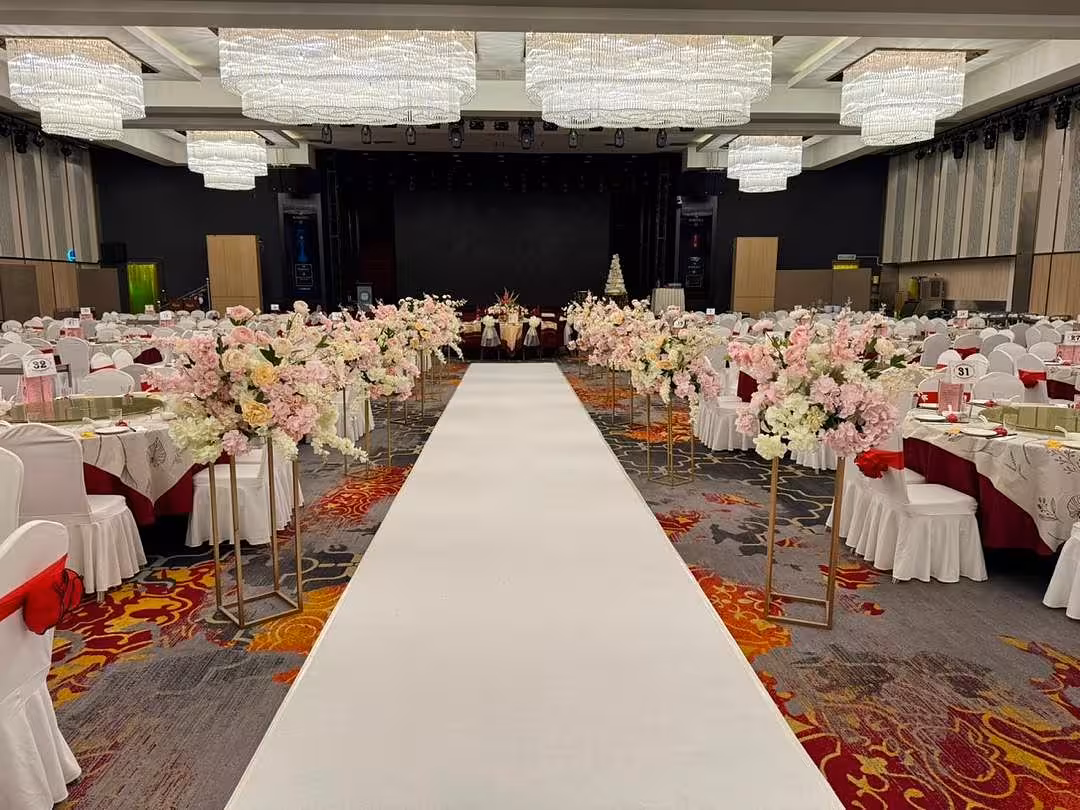 Gold Iron Decorative Event Stands (80 cm) line a pristine white aisle runner in pairs, each topped with lush arrangements of blush pink, peach, and ivory blooms including roses, hydrangeas, and cherry blossom sprays. The grand ballroom setting is completed with white-draped round dining tables accented in deep red, a patterned carpet, and cascading crystal chandeliers overhead.