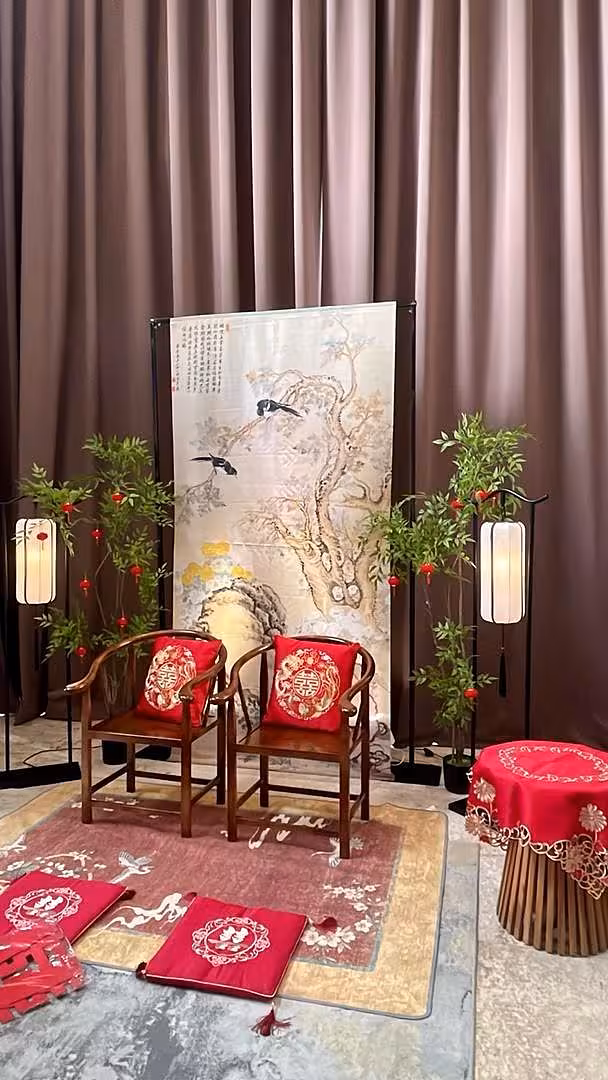 A traditional Chinese tea ceremony vignette styled in rich red and warm wood tones, anchored by a large cypress and bird ink painting hanging cloth framed in a black metal arch stand. Rustic wooden horseshoe chairs dressed with embroidered dragon and phoenix red cushions sit atop a layered oriental carpet, flanked by glowing arch vertical lantern floor lamps, artificial nandina greenery adorned with small red velvet hanging lanterns, and an embroidered red table cloth-covered side stool.