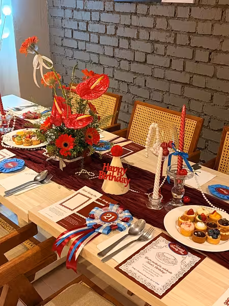 Red anthurium and gerbera centrepiece on a burgundy table runner with glass taper candle holders, birthday props, and place settings
