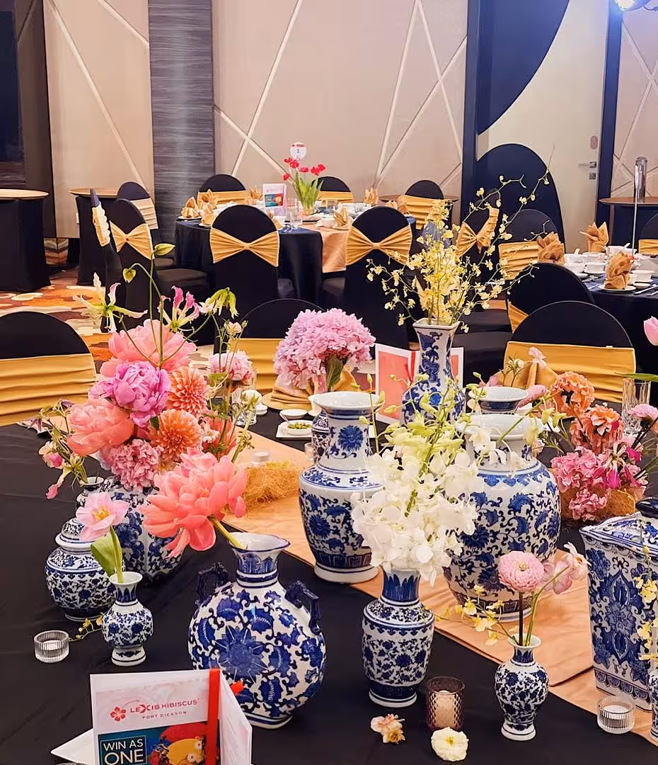 Cluster of mini oriental blue floral porcelain vases in varied shapes filled with pink peonies, dahlias, white orchids, and yellow blossoms on a black banquet table