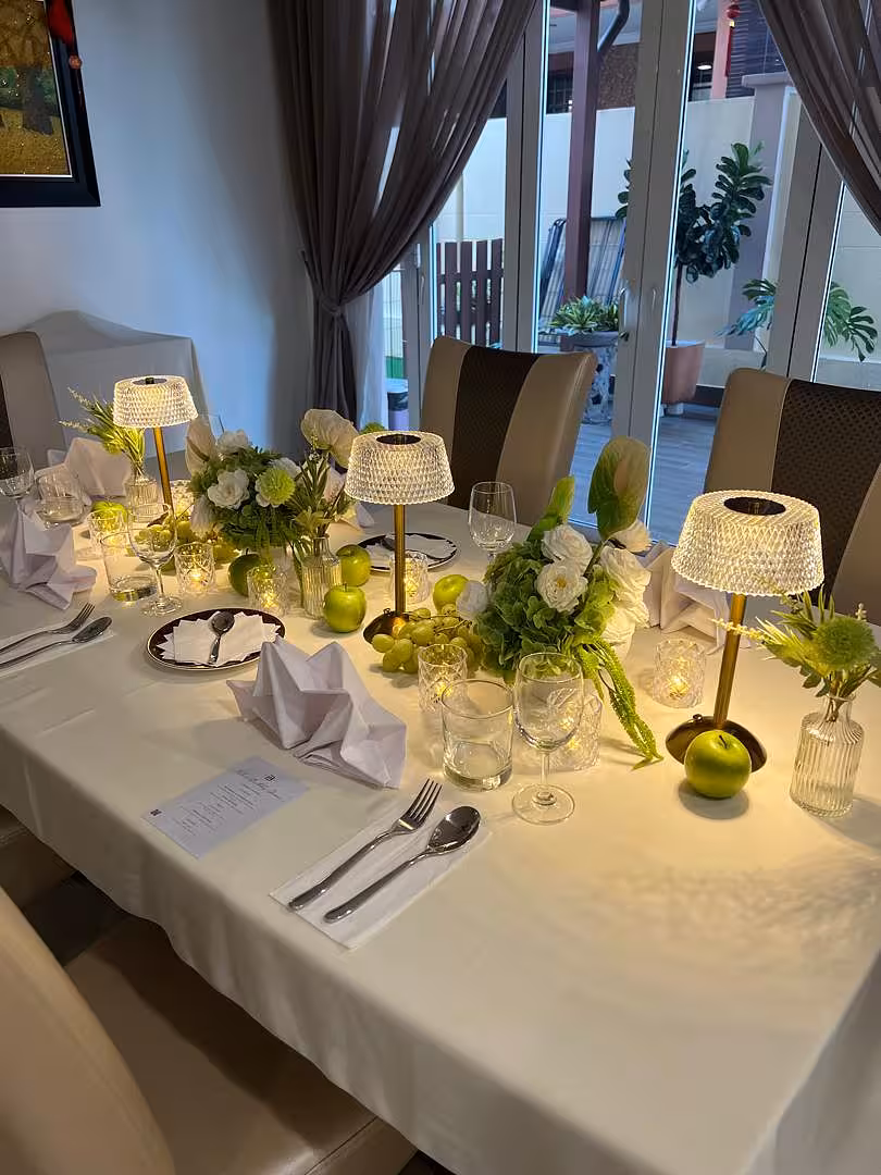 A crisp white tablecloth sets the base for a lush green-and-white tablescape, where Crystal Shade Portable Dining Table Lamps cast a warm golden glow alongside clusters of Eden Artificial Table Flowers in Mini Clear Glass Vases. LED Crystal Effect Tealight Candle Cups flicker softly between place settings, while green apples and grapes echo the fresh botanical palette.