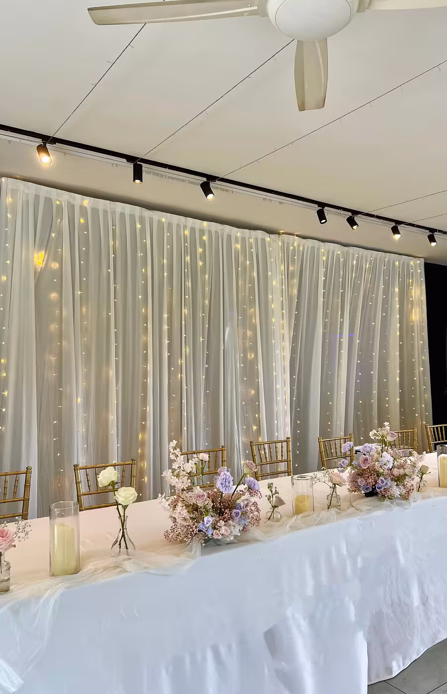 A billowing white sheer fabric curtain backdrop is layered with warm white LED fairy lights, casting a soft, starlit glow across the sweetheart table. The table is dressed in white linen with a cascading tulle runner, pillar candles, and lush pastel floral arrangements in blush, lilac, and dusty rose.