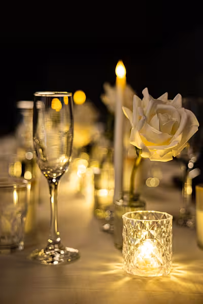 Crystal LED tealight candle cup glowing beside white artificial curl-edge rose bouquet and champagne flutes on a candlelit table
