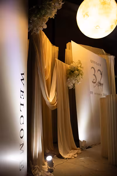 Champagne stretchable drape backdrop with white artificial flowers, a personalised birthday signage panel, and a glowing moon pendant light