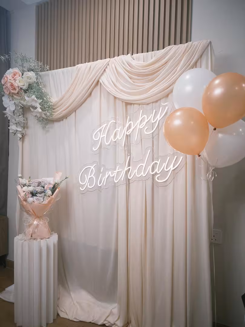 A soft champagne stretchable drape backdrop is elegantly swagged at the top, anchored by a cluster of dreamy pink artificial wall-hanging flowers on one side and peach and white balloons on the other. A glowing white Happy Birthday LED neon sign takes centre stage, while a wrapped floral bouquet rests on a white origami cylindrical pleated dessert stand below.