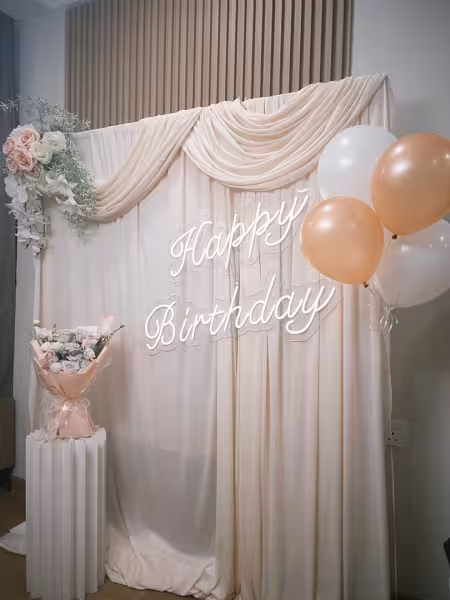 Champagne drape backdrop with Happy Birthday neon sign, pink artificial wall flowers, peach and white balloons, and a floral bouquet on a white cylindrical stand