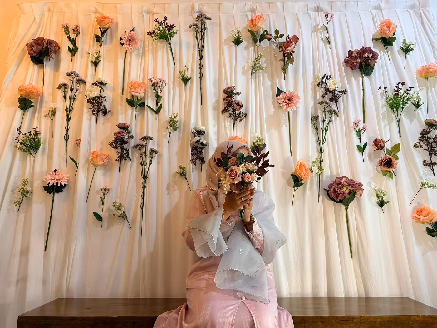The Wedding Fabric Joyous White Celebration Curtain forms a soft, flowing backdrop layered with individually placed artificial blooms in peach, mauve, dusty rose, and cream, interspersed with lush green foliage. The scattered floral arrangement creates a whimsical, garden-inspired aesthetic with a romantic and delicate mood.
