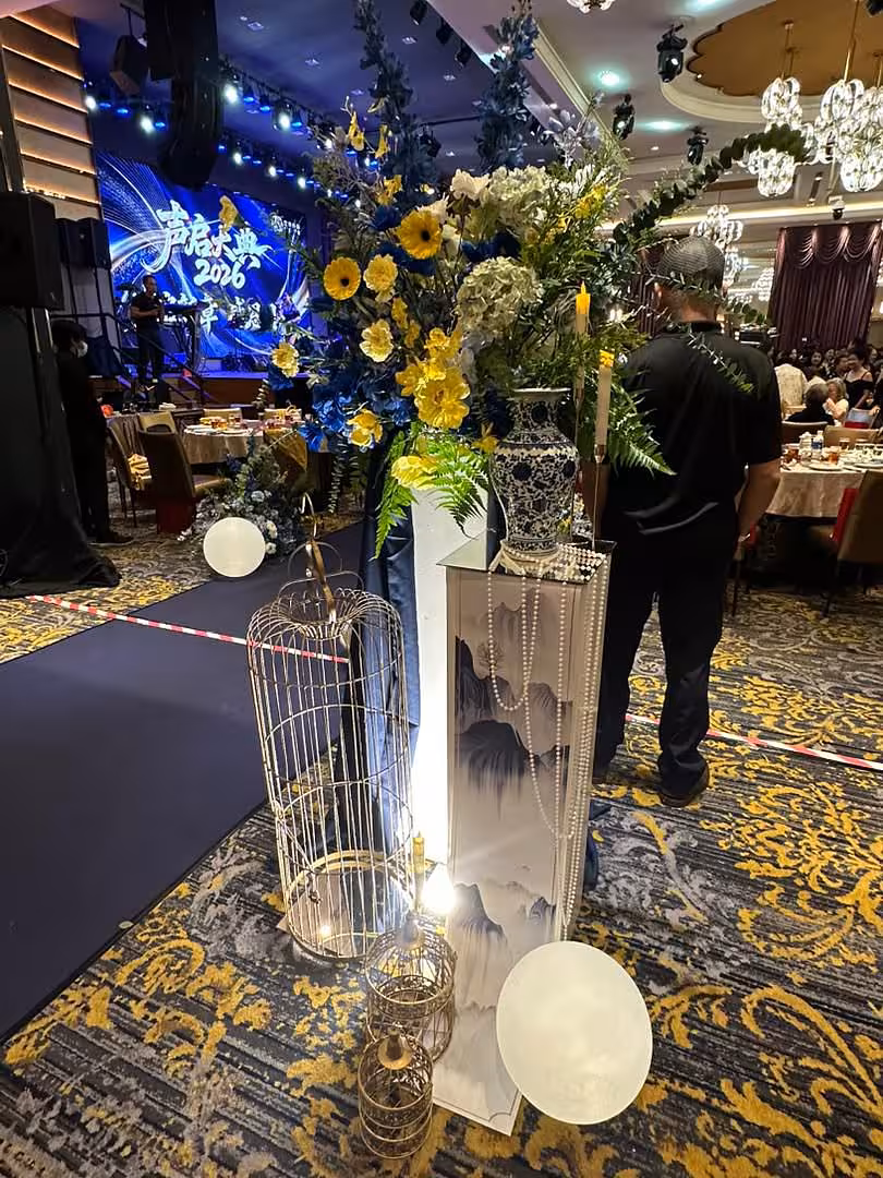 A tall ink-wash landscape pedestal displays a lush arrangement of navy blue and yellow blooms in a blue-and-white porcelain vase, draped with pearl strands. Antique-style gold birdcage lanterns and softly glowing LED ball stone sphere lights anchor the styling along the event aisle.