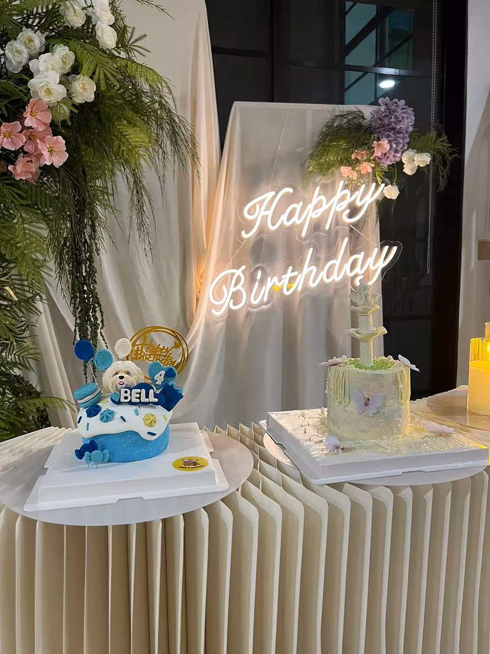 A softly gathered champagne fabric drape forms a dreamy backdrop, anchored by a glowing white Happy Birthday LED neon sign. Sweet Forest artificial wall flowers in white, pink, and lavender with lush trailing greenery frame the top corners, while a structured pleated cream table below completes the elegant, garden-inspired setup.
