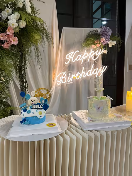 Champagne drape backdrop with glowing Happy Birthday neon sign, sweet forest floral wall clusters, and a pleated cake table