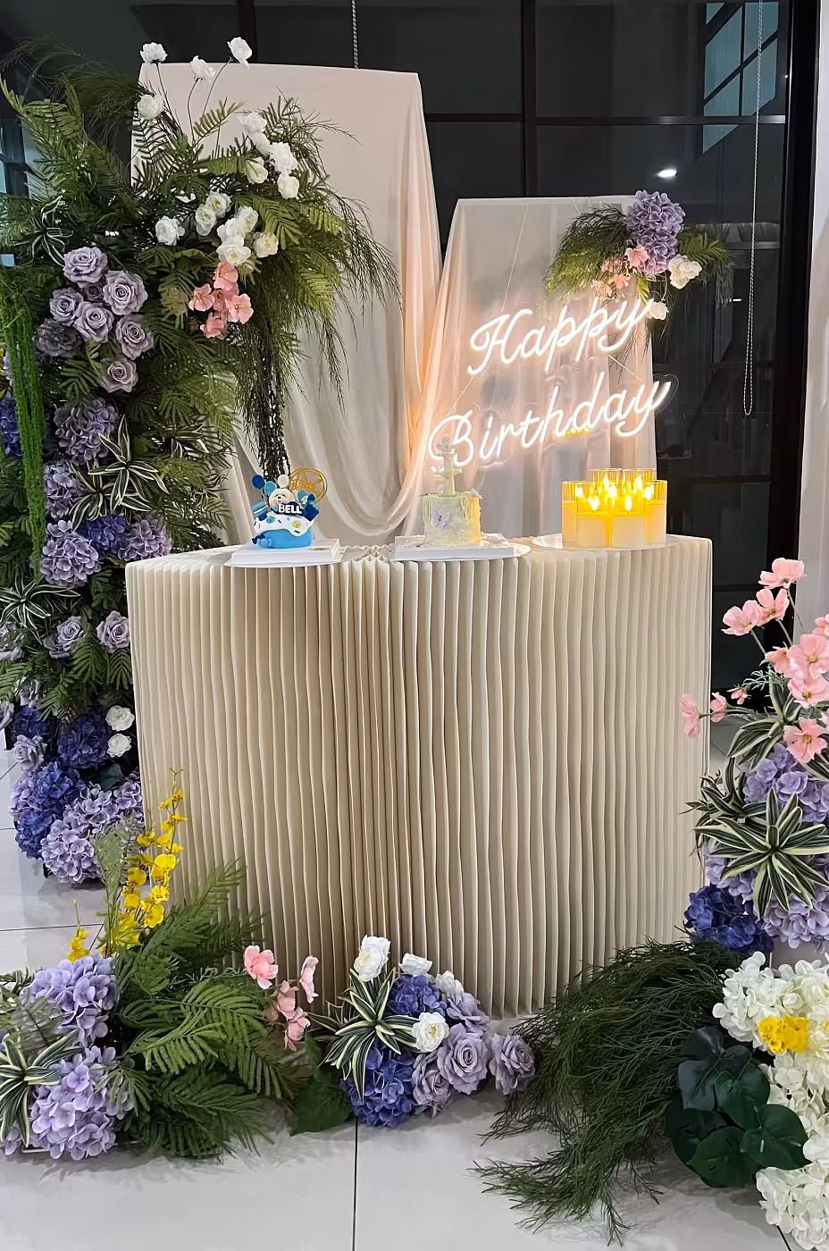 An accordion-pleated Origami Dessert Table sits centre-stage beneath a warm-glowing Happy Birthday LED Neon Light Signage, flanked by lush Sweet Forest Artificial Flower Arches bursting with purple roses, hydrangeas, pink blooms, and tropical greenery. Matching floor and table flowers extend the enchanted garden mood, while a trio of LED Acrylic Candles adds a soft golden glow to the cream draped backdrop.