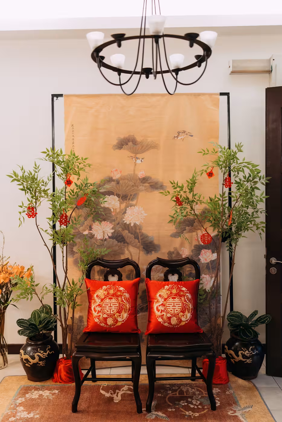 A pair of rustic walnut wooden chairs dressed in vivid red embroidered dragon and phoenix cushions are framed by lush artificial nandina floor ornaments adorned with red double-happiness charms, all set against a warm amber ink painting hanging cloth depicting ducks among lotus blossoms. Black ceramic dragon vases, a floral area rug, and a black square arch complete this elegant, auspicious scene.